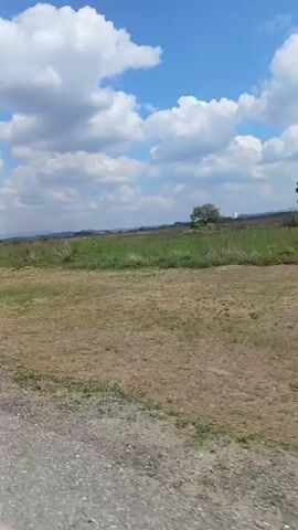 The video depicts a rural landscape on a sunny day with a blue sky dotted with white clouds. A wide, flat field, primarily dry and brown with patches of green, stretches into the distance. The horizon shows a faint outline of hills or mountains. In the foreground is a paved road, likely where the camera is positioned. The camera appears to be moving, creating a panning effect across the landscape. The wind is strong, causing noticeable audio interference.