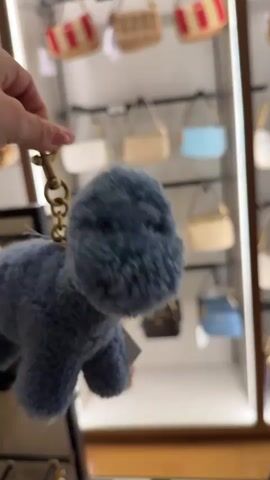 A person's hand holds a fuzzy, blue, plush dinosaur keychain up to the camera. The keychain has a gold-colored clasp and ring. The tag is attached to the dinosaur with a black string. In the background, there is a display case with various small bags or purses.