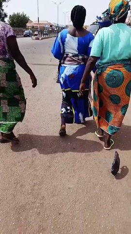 The video shows three women walking away from the camera on a road. They are wearing colorful, patterned fabrics wrapped around their waists, and two of them have matching headwraps. The woman in the middle is wearing a blue and white dress. There are cars and other people in the background. The road is made of a reddish-brown material and there is some debris on the ground.