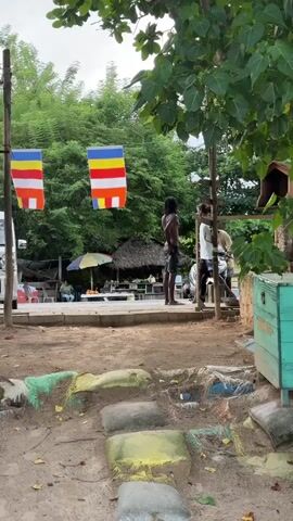 The image depicts a scene in Peleana, Sri Lanka, on an overcast day. In the foreground, a pathway is formed by colorful, rounded objects, possibly repurposed tires, resting on a sandy, dirt ground. Some of these objects are adorned with green and yellow paint. Beyond this path, a paved area is visible, with a few people and a vehicle.

Two prominent Buddhist flags, featuring horizontal stripes of blue, yellow, red, and white, hang from a wooden pole on the left side of the frame. In the middle ground, a thatched-roof structure with an umbrella suggests a casual outdoor seating area, possibly a small eatery or rest stop. Several individuals are present, with two men standing in the center of the frame, one facing away from the camera and the other partially obscured. A person is seated in the background near the thatched structure. Another individual is seen near a parked motorcycle on the right. A light-colored van is visible on the far left. A rustic, turquoise chest is situated on the right side of the image. The background is filled with lush green trees, indicating a tropical environment. The overall atmosphere appears relaxed and informal, typical of a beachside or rural location in Sri Lanka.