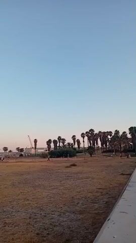A drone surveys an outdoor scene with a wide shot, capturing a beach and a grassy area. The sky is a clear blue with a soft gradient of pink and orange hues near the horizon, indicating either sunrise or sunset. In the background, palm trees line a coastal area, with a long pier or breakwater extending into the water. Several cranes are visible near the coastline, suggesting ongoing construction or industrial activity. A group of people are gathered near the pier and along the beach, some of whom appear to be walking or socializing. The foreground consists of dry, yellowish grass and sand, with some wooden posts and ropes marking a pathway or boundary. The drone then pans to reveal a more densely populated area further along the beach, where a large crowd has gathered. Many individuals are standing in groups, some closer to structures that appear to be businesses or vendors. The overall atmosphere seems to be one of public gathering or an event taking place by the sea.