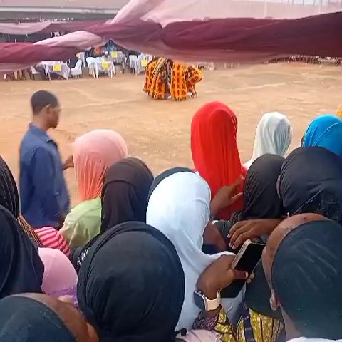 A group of people, mostly women in hijabs, is seen from behind, looking towards an open field where a cultural performance is taking place. In the background, there are canopies with tables and chairs. The ground is made of dirt.