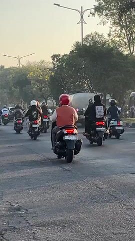 The video shows a street scene with a group of motorcyclists riding in the same direction on a paved road. The motorcyclists are of varying ages and genders, and they are all wearing helmets. Some of the motorcyclists are carrying passengers or cargo. The road appears to be in a city or suburban area, with trees and buildings visible in the background. The sky is clear and the sun appears to be low in the sky, suggesting it is either early morning or late afternoon. The overall impression is of a busy street with traffic.