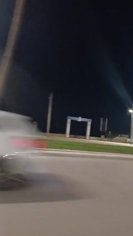 At night, a car passes along a road with streetlights and silhouettes of people. The scene is dark, lit mainly by streetlights and vehicle headlights. There are silhouettes of palm trees and structures like archways. The camera appears to be moving along the road, possibly mounted on a vehicle.
