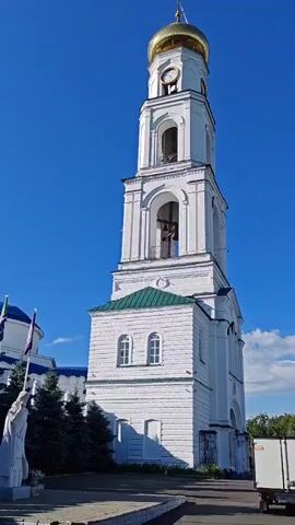 The video shows a tall, white bell tower with a gold dome against a clear blue sky. The tower has multiple levels with arched openings and a clock face near the top. A bell is visible inside one of the openings. At the base of the tower, there is a statue of a robed figure on the left side and some evergreen trees. The ground in front of the tower appears to be paved. A person is walking into the frame from the right at the end of the video.