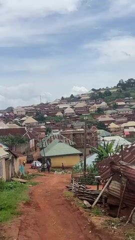 The video shows a hilly landscape filled with numerous buildings, many with colorful corrugated metal roofs. The buildings are clustered together, forming a village or town. In the foreground, a dirt path leads up a slight incline. Several people are visible on the path, some standing and others walking. A motorcycle is parked near the path, and a car is visible further up. Trees and vegetation are scattered throughout the scene, particularly on the hillsides. The sky is partly cloudy, with patches of blue visible. Some of the buildings appear to be under construction, with exposed brickwork and scaffolding. The overall impression is of a vibrant, populated community situated in a natural, somewhat rugged environment.