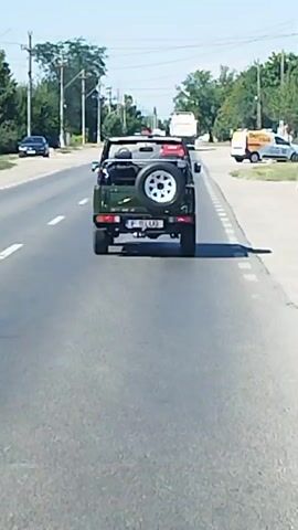 A dark green off-road vehicle with a spare tire mounted on the back drives on a paved road. The vehicle has its top down. A license plate is visible on the rear of the vehicle. The road has white lane markings and a white dashed line separating the lanes. On the right side of the road, there is a curb and a sidewalk. Trees and utility poles line the sides of the road. The sky is clear and blue, indicating a sunny day. Other vehicles are visible in the distance.