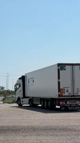 A white Volvo semi-truck with a white trailer is shown from a slightly low angle, moving from left to right across the frame. The truck is on an asphalt road, likely a highway interchange, with a clear blue sky above. The trailer has blue lettering that reads "BESTRAVEL TRANSPORTE NAZIONALE E INTERNAZIONALE" and an address in Castel San Pietro Terme. The truck has multiple axles and appears to be in motion. The lighting suggests it is daytime, possibly late morning or early afternoon.