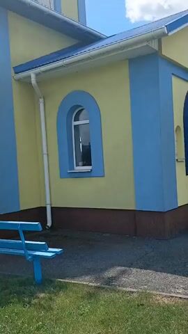The video shows the exterior of a yellow and blue church under a partly cloudy sky. The church has a blue roof and arched windows with white frames. A cross is visible on top of the church's dome. To the right of the church, a small wooden stable structure is set up with figures depicting a nativity scene. The ground is covered with grass and asphalt. There is a blue bench in front of the church.