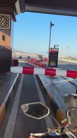 The video captures a first-person perspective from a motorcycle approaching a toll booth. A red and white striped barrier arm is partially lowered, blocking the lane. A traffic light above the lane displays a red signal. In the background, several motorcycles and a yellow bus are visible queuing at other lanes, which appear to have green lights and open barriers. The toll booth structure features a decorative patterned panel on the left. The sky is clear and blue, indicating daytime. The motorcycle's rearview mirror is visible in the foreground.