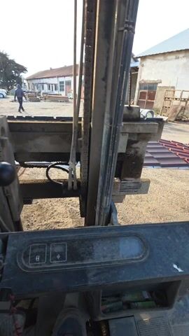 The video shows a first-person view from the operator's seat of a forklift. The forklift mast is prominent in the foreground, with chains and hydraulic lines visible. Outside the forklift, the ground is dirt. A person walks away from the forklift towards a building. There are other buildings, parked cars, and some stacks of roofing tiles in the background. The operator's hand is visible on the steering wheel, adjusting the wheel.