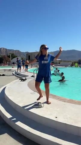 A young blonde woman wearing a blue t-shirt with "STAFF" printed on it and blue shorts is dancing on the edge of a swimming pool. She is barefoot and has sunglasses on. There are people in the pool and other staff members in the background. The sky is clear and blue.