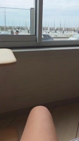 The video shows a view through a window looking out onto a marina. The foreground is dominated by a brown wall and the edge of a light-colored table. In the middle ground, outside the window, there is a street with several cars. A black mini cooper is driving down the street, and a silver car is parked on the right. Beyond the street is the marina, filled with numerous sailboats. There are a few people visible on the dock between the street and the boats. The sky is overcast. The camera is positioned as if someone is sitting at the table, looking out the window.