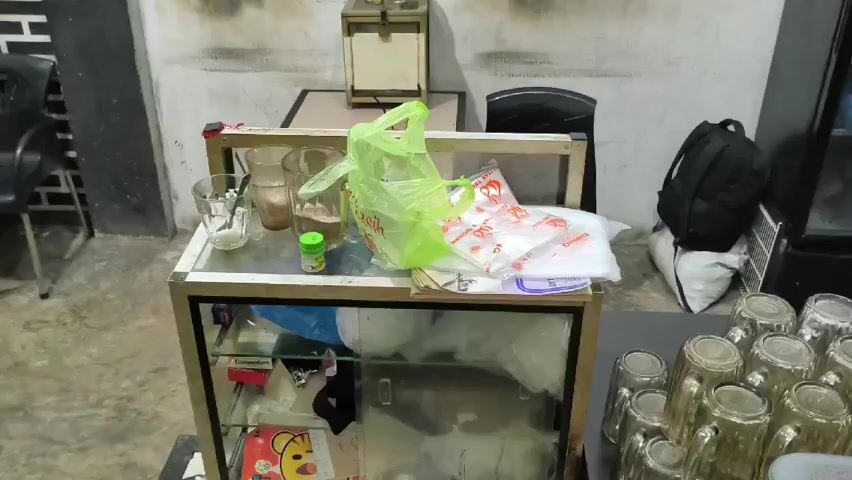 The video shows a small, indoor cafe setting. A glass-topped counter holds several drinking glasses, some with a dark substance inside. A green plastic bag, a small container, and plastic bags sit on the counter. Underneath the counter, there's a box of cigarettes, a notebook, and various items. To the right of the counter, several glass mugs are arranged on a table. A black backpack is resting on a white bag next to the mugs. Behind the counter, a small, wooden box sits on a table, with two glass jars on top. A black chair is positioned behind the table. The walls are white with some stains.