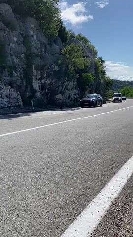 The video captures a view of a two-lane asphalt road in Gornja Brova, Montenegro, on a bright, sunny day with scattered clouds. The road features clear white lane markings and a solid white line defining the shoulder. To the left of the road, a steep, rocky hillside covered with sparse green trees and bushes rises. In the distance, green hills and a partially cloudy blue sky are visible.

The scene depicts traffic moving along this rural road. At the beginning, a dark SUV is parked on the left shoulder of the road. A white truck, possibly a tanker or utility vehicle, drives past the camera moving away, followed by a dark sedan. The previously parked dark SUV then pulls away and drives down the road, and another dark SUV is seen driving towards the camera in the distance. The license plate of the initially parked dark SUV briefly shows "PG," which is a common prefix for Podgorica, Montenegro. No people are visible in the video.