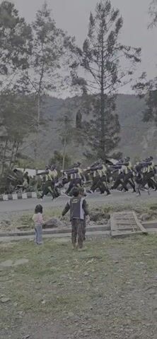 A group of people in matching uniforms, some wearing yellow vests, are marching in formation on a paved road. In the foreground, two children are watching the procession. The environment appears to be outdoors with trees and hills in the background. The sky is overcast.