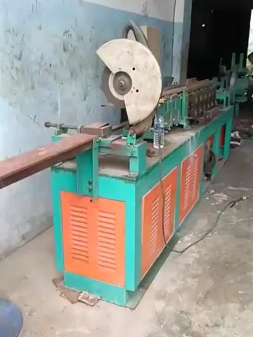 A metal processing machine is shown in operation. The machine is green and orange and appears to be in a workshop. A young man, wearing a black t-shirt, is standing next to the machine, observing it. He is wearing gloves. A metal bar is being fed into the machine. The machine has a cutting wheel on the left side and a series of rollers in the center, suggesting it is used for shaping metal. There is a water bottle on top of the machine. The background shows a concrete wall with some handprints.