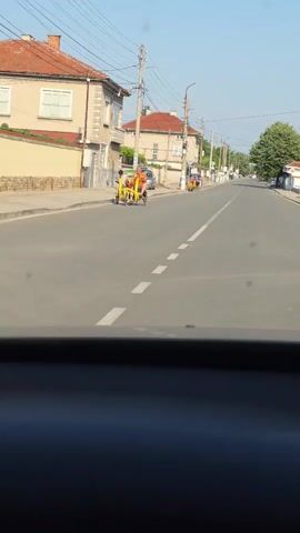 The video, shot from inside a moving vehicle, shows a residential street on a sunny day. A dark dashboard is visible at the bottom of the frame. The asphalt road has a broken white center line and a solid white line marking the right edge. On the left side of the road, two men are pedaling a bright yellow four-wheeled recumbent bicycle, moving towards the camera. One man wears an orange shirt, the other a black shirt. Further down on the left, two individuals stand near a green trash can and a utility pole, while a silver sedan is parked nearby. Another person in a black shirt stands further along the sidewalk on the left. Buildings with red tile roofs line both sides of the street, with utility poles and overhead wires running parallel to the road. In the distance on the right, another yellow four-wheeled pedal vehicle is visible. Trees are present on both sides of the street.