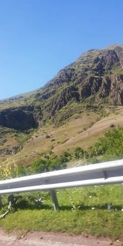 The video captures a view of a mountainous landscape with a clear blue sky. The mountain is covered in lush green vegetation and appears rugged and rocky. In the foreground, a white guardrail runs along the side, with some greenery and a few small white flowers visible near it. The camera angle is tilted, making the scene appear sideways. The video appears to be taken from a moving vehicle, likely along a road, given the presence of the guardrail and the consistent angle of view. There are no people or animals visible in the frame. The overall impression is of a natural, scenic environment.