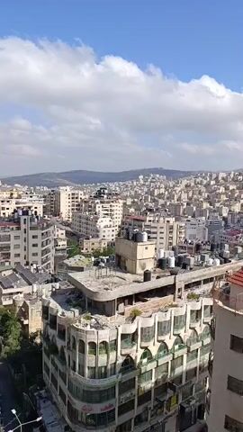The video pans across a dense urban landscape with buildings of varying heights and architectural styles. The buildings are primarily made of concrete and stone, with many featuring balconies and rooftop water tanks. A hillside covered in more buildings is visible in the background, suggesting a city built on uneven terrain. The sky is blue with scattered white clouds, indicating fair weather. There are no people or vehicles clearly visible in the footage.