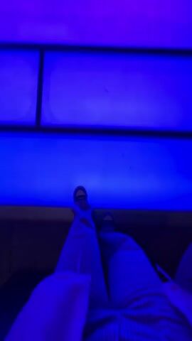The video shows a person walking on a light-up floor that changes color as they move forward. The floor is made up of rectangular tiles. The color changes from blue to purple to red to orange to green. The person is wearing pants with a plaid pattern and white sneakers. The surrounding environment appears to be a hallway or walkway with walls on either side. Small lights can be seen in the ceiling.