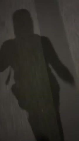 A shadowy figure's silhouette is cast on a dark, wet street. The figure runs with arms swinging and legs pumping, their shadow appearing to move quickly over the reflective surface. The only discernible color is a small blue object visible at the bottom of the frame, likely the tip of a shoe. The street has a dark, reflective stripe down the center.