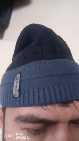 A close-up shot focuses on a person wearing a two-tone blue and black beanie. The beanie has a small, rectangular label on the blue portion. The person's eyes and eyebrows are visible. The background is a plain white wall. Part of a dark blue jacket is visible in the upper left corner. The video is recorded using a TECNO SPARK 9 Pro device.