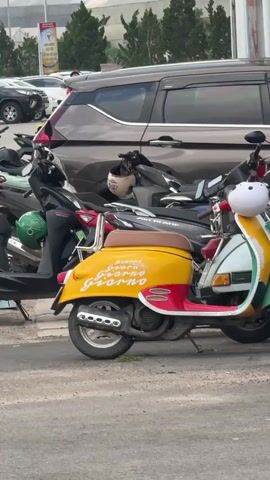 The video shows a parking area with a row of parked scooters, including a yellow, red, and white Giorno scooter with "Giorno Giorno Giorno" written on its side. Behind the scooters is a dark gray minivan. Two young men in casual clothes are walking near the scooters. The parking area is in front of a tall building with white columns. In the background are more buildings and some trees. The sky is overcast.