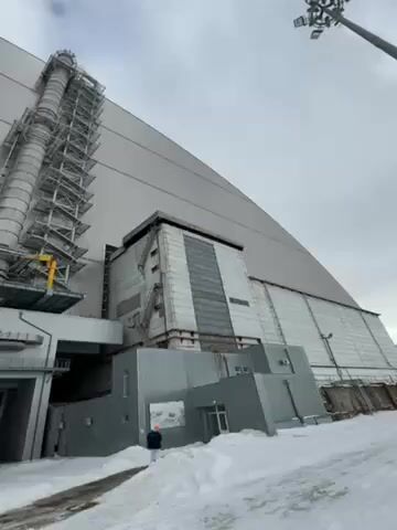The video shows the New Safe Confinement (NSC), a large arch-shaped structure built over the Chernobyl Nuclear Power Plant's damaged Reactor Number 4. The structure is massive, gray in color, and dominates the frame. To the left of the NSC, there is a tall vertical structure, potentially a ventilation or exhaust system, covered in scaffolding. Adjacent to the NSC, are various smaller buildings or annexes, also gray, and showing signs of wear and aging. Snow covers the ground, suggesting a cold climate. A person wearing a dark jacket is standing near the base of one of the buildings, close to an entrance. The sky is overcast and gray. The camera is looking up at an angle, emphasizing the scale of the NSC.