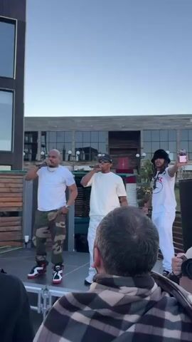 Three men are on a stage performing. The man on the left wears a white t-shirt and camouflage pants with red and black sneakers. The man in the middle wears a white t-shirt, a black baseball cap, sunglasses, and white camouflage pants with white sneakers. The man on the right wears a white t-shirt and white pants with a black hat. He is holding a phone in his right hand. They are all holding microphones and appear to be rapping or singing. There is a crowd of people in front of the stage, some of whom are visible in the foreground. The background shows a building with windows and a wooden fence. The sky is clear and appears to be daytime.