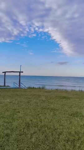 The video opens with a wide shot of a grassy area in front of a calm ocean under a partly cloudy sky. A wooden structure with blue railings is visible on the left, leading down a set of stairs. As the camera pans to the right, a blue bench or structure appears on the grass near the waterline. The scene is serene and peaceful, depicting a natural landscape at what appears to be daytime. There are no people or animals visible in the frame.