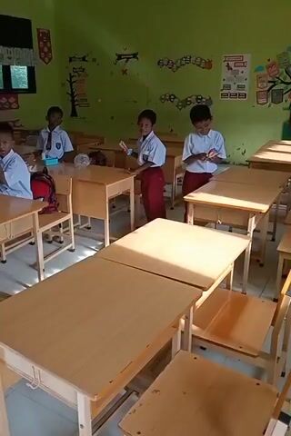 The video shows the interior of a classroom with several young boys dressed in school uniforms, consisting of white shirts and red pants. The room is decorated with colorful posters and drawings on the light green walls. There are rows of wooden desks and chairs arranged in the classroom. The boys appear to be engaged in some activity, possibly a lesson or a break. Sunlight streams in through a window, illuminating the desks and the floor. The overall atmosphere seems calm and academic.
