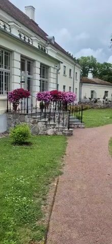 A panning video shows a large building with white walls and large windows, surrounded by a manicured lawn and trees. Red and pink flowers in pots are arranged along a stone staircase leading to the building's entrance. A gravel path winds through the lawn, leading past a white bench and a small fountain. The sky is overcast.