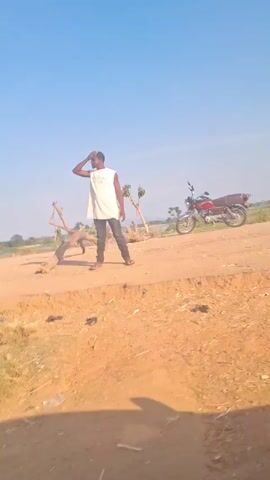 The video shows a man standing near a dirt road, with a motorcycle parked nearby. The man is wearing a white tank top and dark pants and is shielding his eyes. The motorcycle is red and black. The road is unpaved and dusty. In the background, there is a flat landscape with sparse vegetation, and a body of water is visible in the distance. The sky is blue with a few clouds. The camera moves forward along the dirt road, showing more of the landscape.