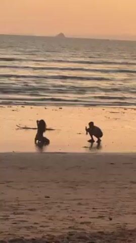 A person is taking a photo of another person on a beach at sunset. Both people are silhouetted. The person posing is kneeling in the wet sand at the edge of the water. The person taking the photo is crouched down and holding a phone. The beach is sandy, and the water is calm. There is a small island visible in the distance. The sky is a warm orange color. There is some debris on the sand.