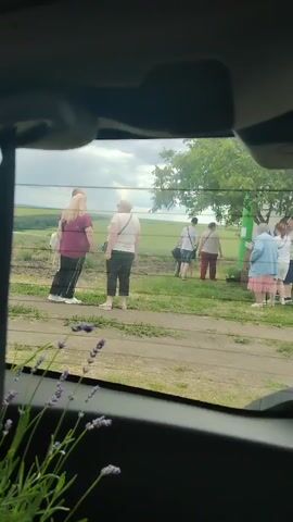 A group of people are gathered outside near a small building. The perspective is from inside a vehicle, looking through the rear window, with the edge of the windshield and part of a sun visor visible at the top. Lavender flowers are in the foreground, partially obscuring the view. The people are standing on a dirt path or grassy area. Some are facing away from the camera, while others are looking towards a small, blue and white kiosk-like structure. The kiosk has displays and shelves visible through its windows, suggesting it might be a shop or concession stand. The overall environment appears to be rural or semi-rural, with fields and trees in the background under a cloudy sky. The individuals are dressed in casual summer clothing. Their actions are not clearly discernible, but they appear to be milling around or perhaps queuing.