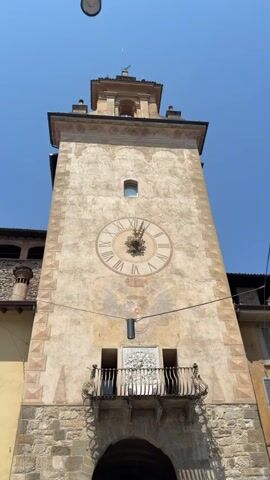 The video shows a low-angle shot of a clock tower against a bright blue sky. The tower has a large, round clock face with Roman numerals and ornate hands. Below the clock face is a small balcony with a wrought-iron railing and intricate decorative carvings. The tower is made of stone and plaster, with faded painted details. A black cylinder, possibly a light or speaker, is mounted below the clock face. The tower appears to be part of a building with arched entrances at the base. The camera is stationary.