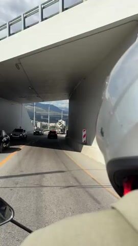 The video begins inside a tunnel. A motorcycle is traveling through the tunnel, with the view from behind the rider's helmet. Other vehicles are visible on the road ahead, including cars and a large truck. As the motorcycle exits the tunnel, a city and mountains are visible in the distance. The road is paved and has yellow lane markings. There are power lines and streetlights along the side of the road. The sky is blue with scattered white clouds. The motorcycle continues along the road, which appears to be an elevated highway or bridge.