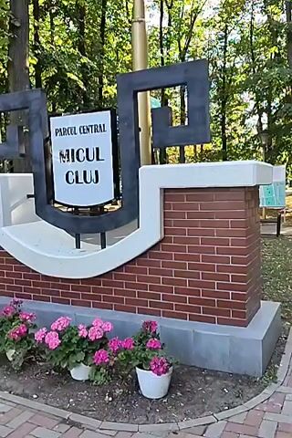 The video shows a sign for "PARCUL CENTRAL MICUL CLUJ" in a park setting. The sign is large and made of metal and brick. There are pink flowers in pots in front of the sign. The park is full of trees and greenery. The video is filmed from a low angle, looking up at the sign. There are no people visible in the video.
