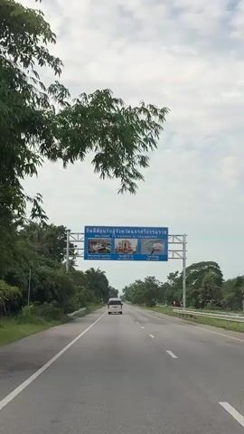 A white van drives down a highway under a cloudy sky. A large blue sign spans the highway with images and text in Thai and English. The sign reads "WELCOME TO NAKHON SI THAMMARAT" and features pictures of a temple, a statue, and a pink dolphin. The highway is lined with green trees and foliage. The camera is positioned from inside a vehicle looking forward.
