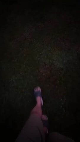 This is a first-person perspective video, filmed at ground level and looking down at the subject's feet and legs as they walk. The subject is wearing dark shorts and dark blue slip-on sandals. They are walking on a grassy area. The lighting suggests it is either dusk or night, with the grass appearing dark and the only illumination coming from an unseen light source that casts a faint glow on the grass ahead. The motion blur indicates the person is walking at a moderate pace. The setting appears to be outdoors, likely in a park or backyard given the grass. No text is visible in the video. The overall mood is somewhat subdued due to the low light conditions. The location context indicates this was filmed in Lapės, Lithuania.