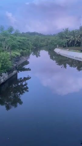 The video shows a view of a dark, calm river or canal flowing through a lush green landscape with trees and vegetation on both sides. The sky above is cloudy and appears to be either dawn or dusk, with soft, diffused light. The water reflects the sky and the surrounding scenery, creating a mirror-like effect. The banks of the waterway are visible, with one side appearing more natural and overgrown with trees, while the other side has a man-made, concrete embankment. In the distance, hills or elevated terrain covered in trees are visible. There are no people or active elements in the scene; it is a static, natural landscape.