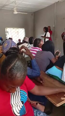 The video shows a classroom scene with rows of desks occupied by various people. In the foreground, a person in a red shirt with a black and white pattern is seated, and only the back of their head is visible. Other individuals can be seen sitting at the desks, some of them looking at their phones or resting their heads in their hands. At the front of the room, a woman wearing a white top and a black hijab is holding a document and appears to be addressing the room. Another woman in a red shirt is standing nearby, seemingly assisting or co-presenting. There is a ceiling fan visible above, and a window with a barred view to the outside. The room's walls are a muted gray, and the overall atmosphere seems to be of a casual, perhaps educational, setting.