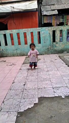 A young child stands on a cracked, tiled surface, moving their arms and legs. They are wearing a light pink t-shirt and patterned pants. Behind them, a weathered wall is painted in teal with vertical red stripes, and a building with a corrugated roof is visible. The tiles underfoot are a mix of pink and gray, showing extensive cracking and damage. The setting appears to be an outdoor space adjacent to some buildings.