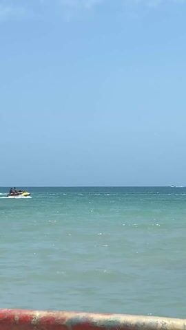 The video shows a beach on a sunny day. The water is a clear turquoise color with small waves. In the distance, a yellow and black jet ski is speeding across the water, leaving a white wake behind it. There is another boat further out in the ocean as well. In the foreground, several people are swimming and playing in the water. There is a rusty metal barrier at the very bottom of the frame.