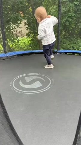 A child walks across a trampoline. The child is wearing a white shirt with pink and red floral patterns, dark blue pants, and white shoes. The trampoline is black with a blue rim and safety net. Trees and foliage are visible in the background.