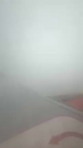 The video shows a plane wing through a window, partially obscured by thick, gray clouds. The plane is flying through overcast skies, and the wing features a red and white design with an arrow indicating direction.