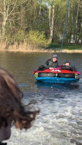 The video shows a boat pulling an inflatable tube on a lake on a sunny day. Two people are riding in the tube, a man and a woman. The man is wearing a black wetsuit and a blue hat, while the woman is wearing a black wetsuit and a pink top. The tube is blue, red, and yellow. The boat is creating a large wake as it moves through the water. In the background, there are trees and a large house.