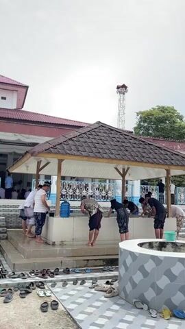The video shows a group of people performing ablutions at a mosque. They are using a communal washing station with several faucets. The people are dressed in traditional Muslim attire, including sarongs and skullcaps. There are also several pairs of shoes and sandals neatly arranged on the ground around the washing area, indicating a place of worship. The background shows the mosque building and a tower with a speaker, suggesting it is a place of prayer and religious gathering. The sky is overcast.