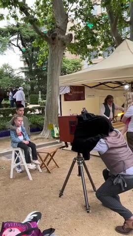 A boy and girl sit on a white stool posing as an older man, dressed as a photographer with a vest and pinstripe pants, crouches to take their picture using an antique box camera with a tripod.  The photographer has his head under a black cloth behind the camera.  The children are sitting in front of a tree in a park or garden setting, with a small wooden folding table next to them. Behind the photographer and to the right is a white tent with a sign in a foreign language and other people gathered. A bag decorated with a cartoon character is in the lower left corner.